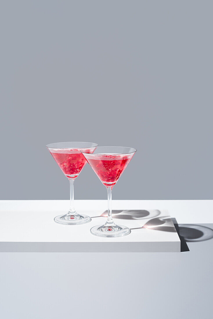 Mit roten Granatapfelcocktails gefüllte Gläser vor einem gedämpften grauen Hintergrund, der einen weichen Schatten wirft