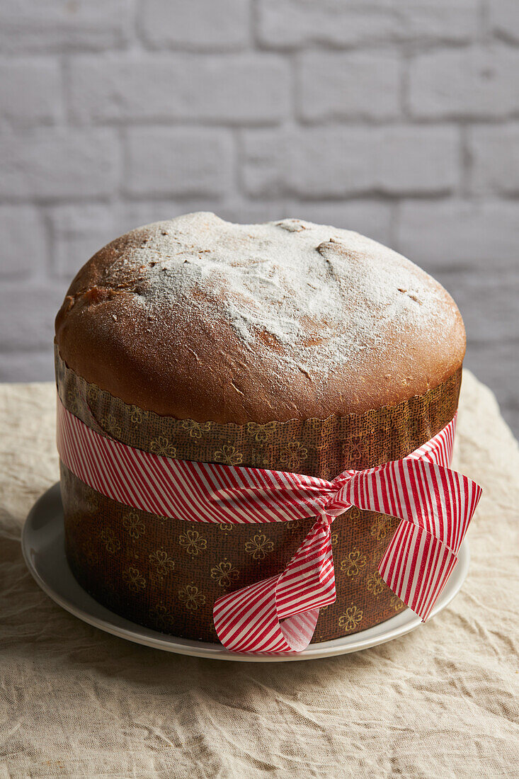 Elevated view of a fresh panettone, dusted with powdered sugar, wrapped with a festive ribbon, set against a brick background