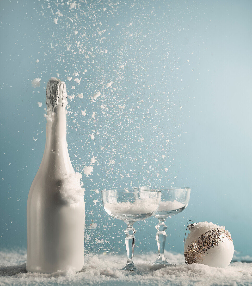 Weiße Champagnerflasche und Gläser mit Weihnachtskugel auf dem Tisch mit Schneefall an blauer Wand Hintergrund. Neujahrskonzept mit Getränk und fallendem Schnee. Frontansicht