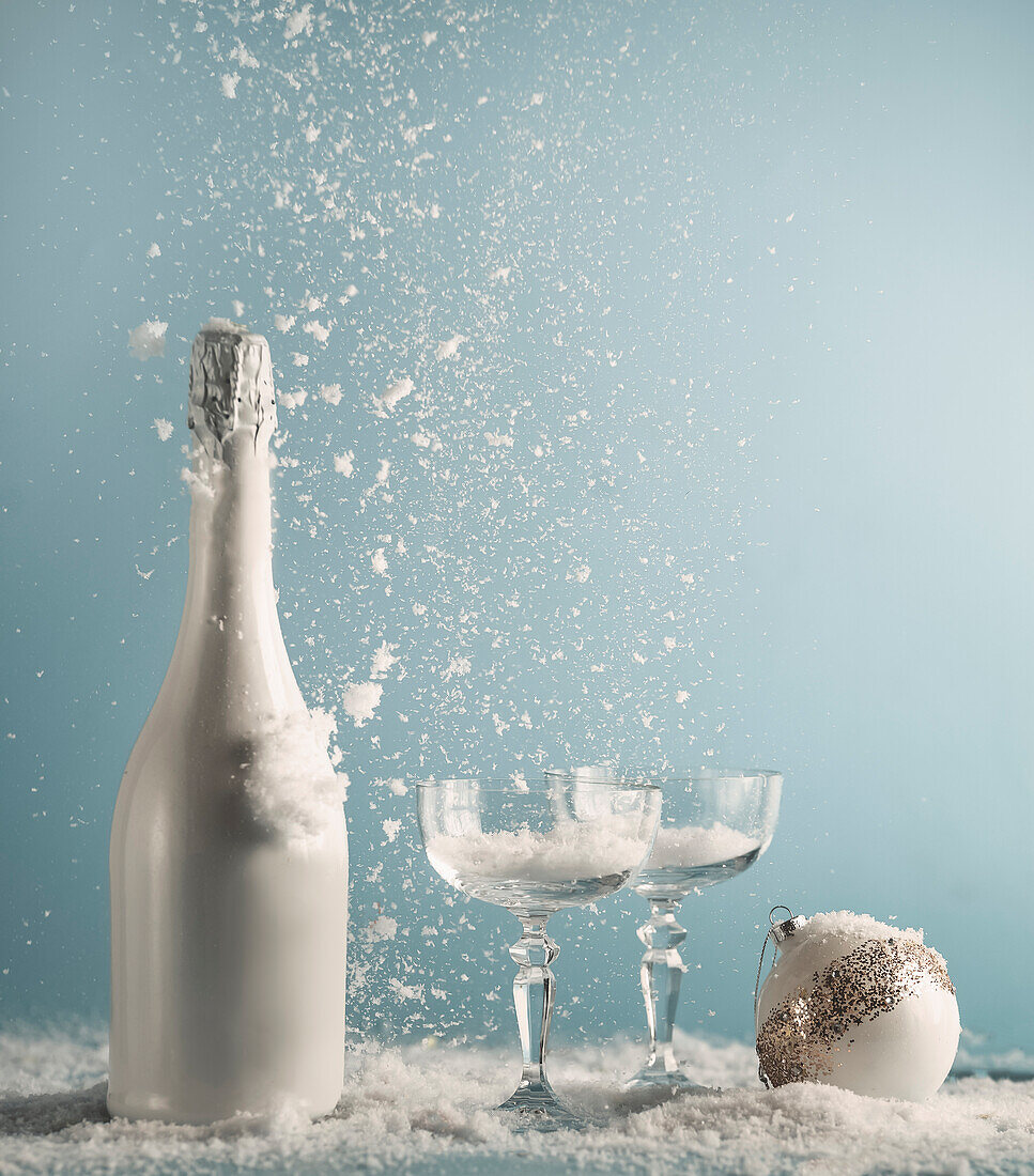 White champagne bottle and glasses with Christmas bauble on table with snowfall at blue wall background. New years concept with drink and falling snow. Front view.