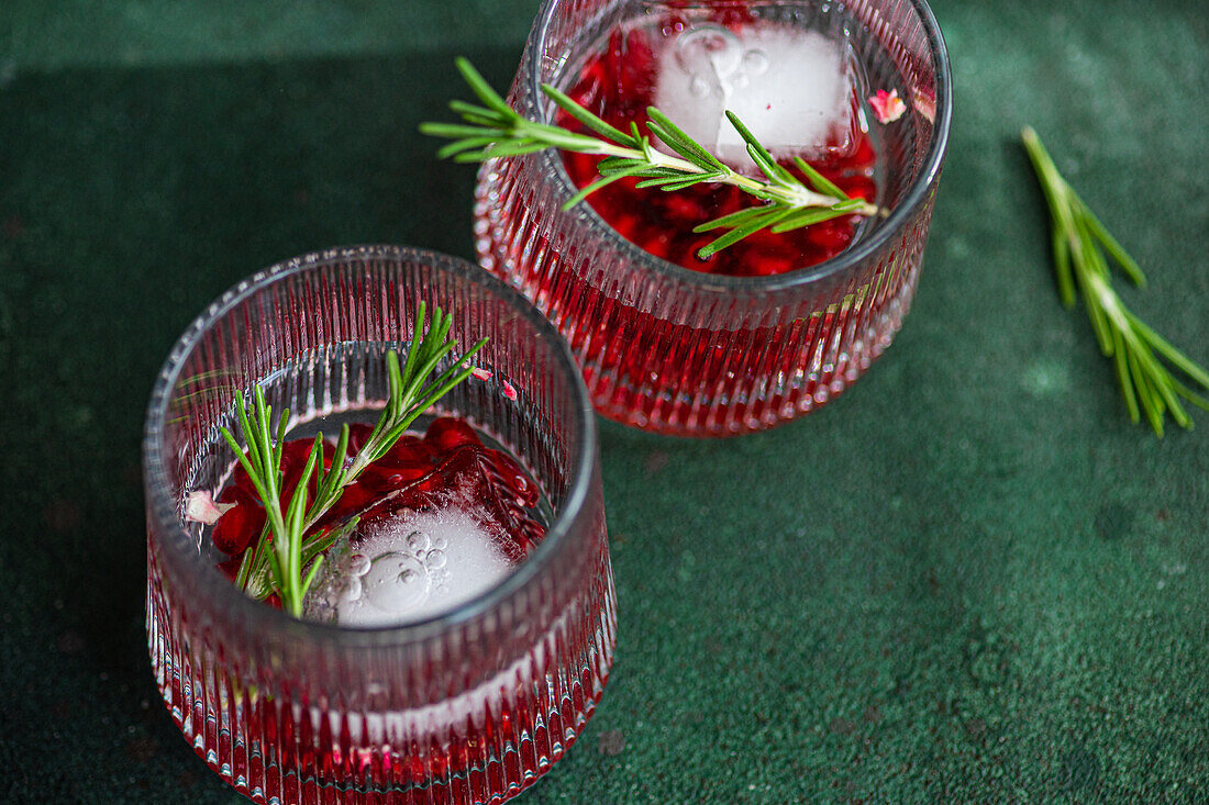 Draufsicht auf zwei Gläser mit Gin-Tonic-Cocktail mit Granatapfelkernen und einem Rosmarinzweig auf einer dunkelgrünen, strukturierten Oberfläche mit einem einzelnen Rosmarinzweig daneben
