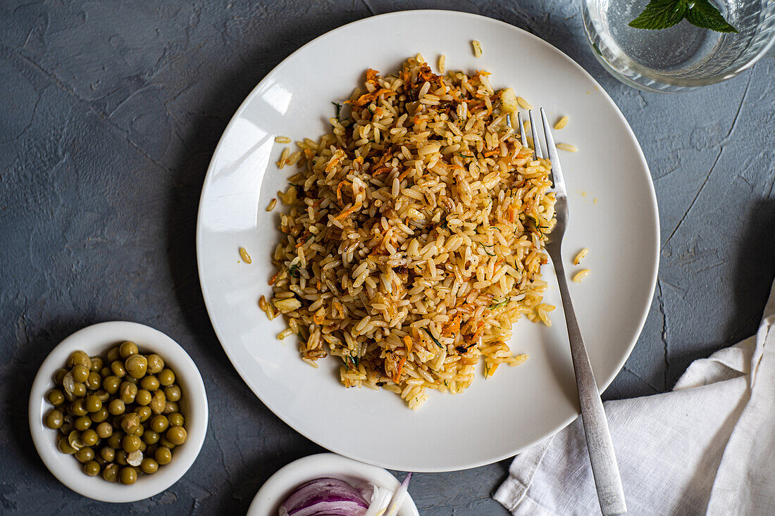 Top view of boiled rice with onion, carrot, bell pepper, green peas and spices on plate with fork near slices of onion and glass of water with mint against gray background