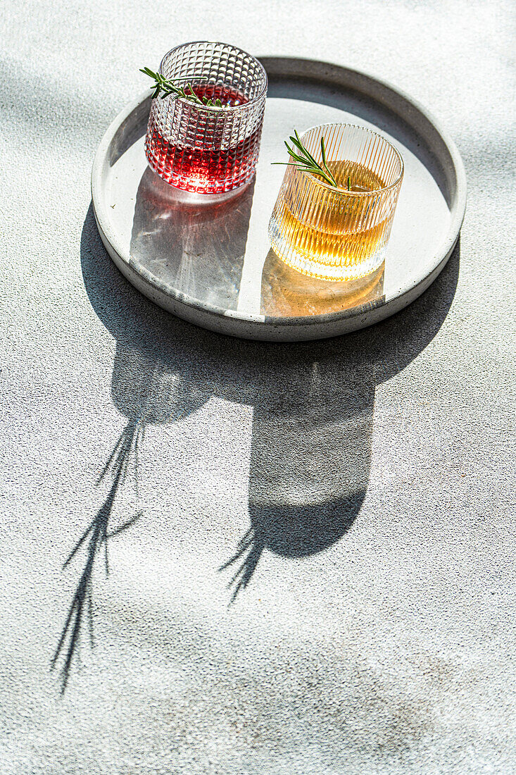 From above of artisanal cocktails with vodka cherry, and apple juice in textured glasses adorned with rosemary on a concrete surface with shadows