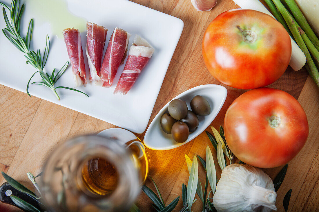 Hochwertiges Bild mit verschiedenen frischen Zutaten, darunter in Scheiben geschnittene Tomaten, iberischer Schinken, Olivenöl, Oliven, Spargel, Knoblauch und Rosmarinzweige, die auf einer Holzfläche angeordnet sind