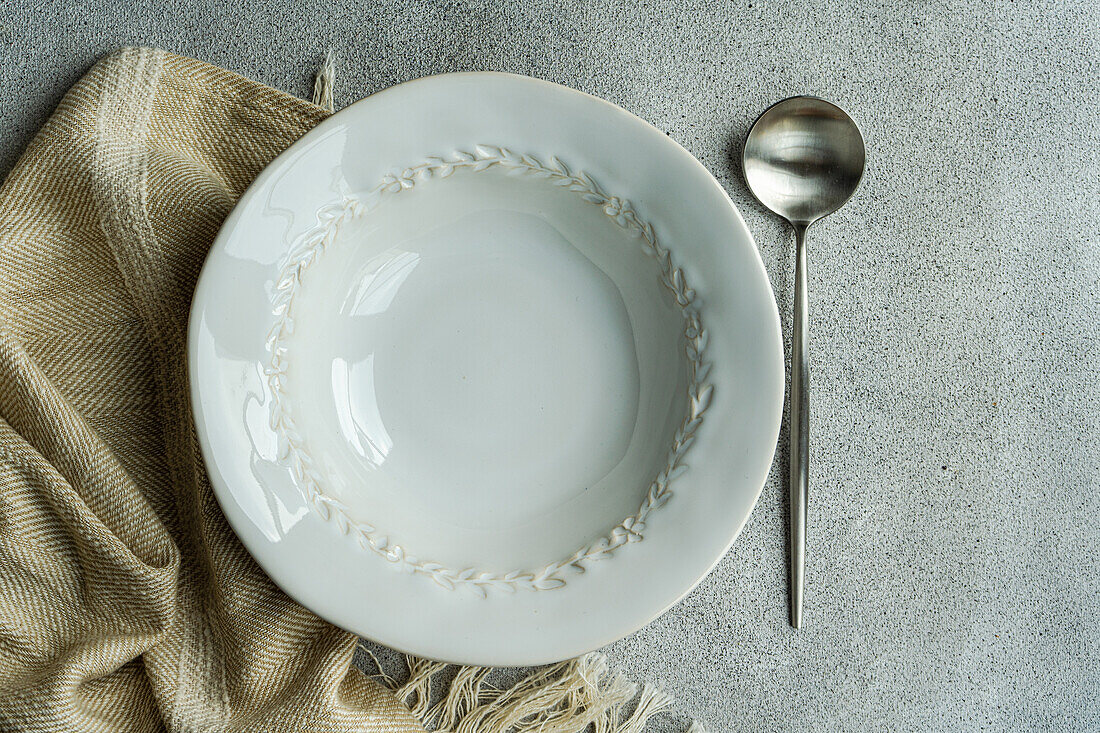 Top view of autumnal table setting with white ceramic bowl between spoon and napkin against gray surface