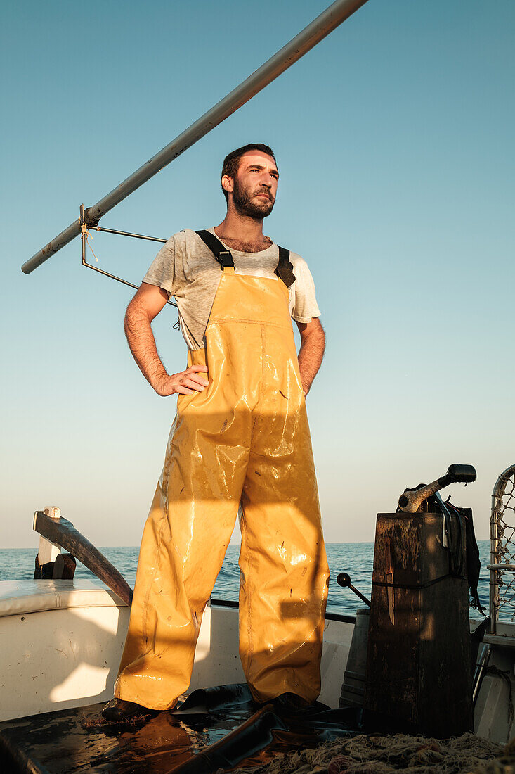 Nachdenklicher männlicher Fischer in Uniform in voller Länge, der die Meereslandschaft bewundert, während er mit einem Netz auf einem Trawler in Soller in der Nähe der Baleareninsel Mallorca fischt