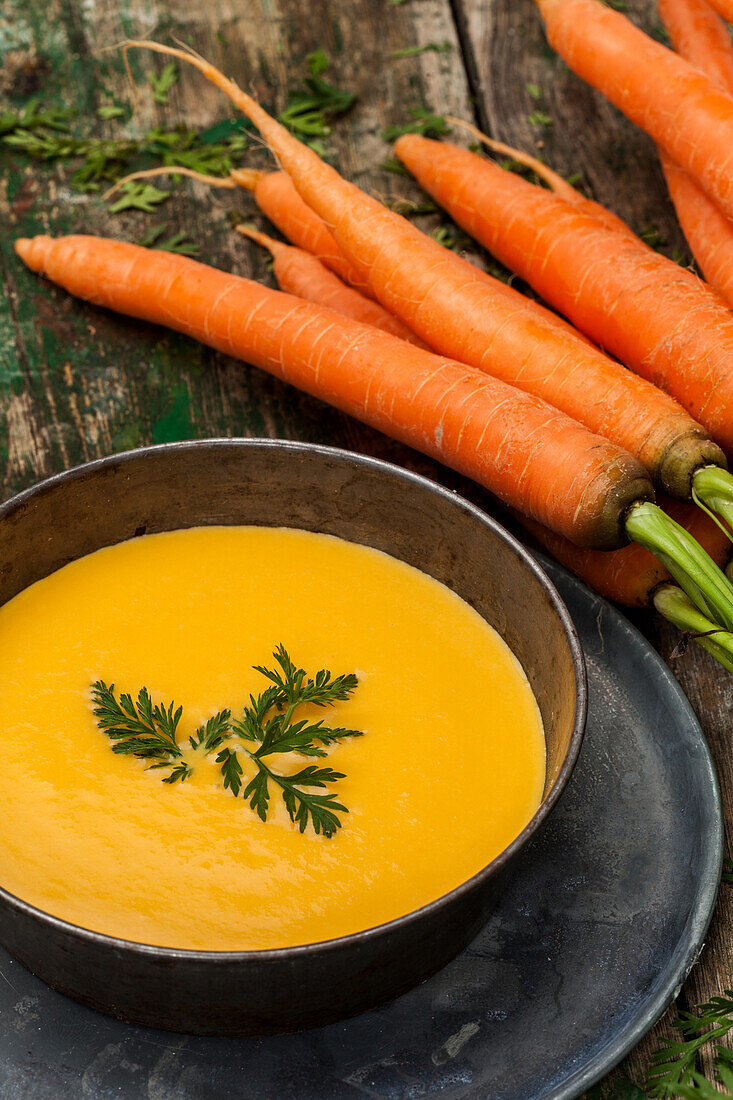Fresh carrot soup in a bowl with whole carrots on a rustic wooden table, garnished with parsley.