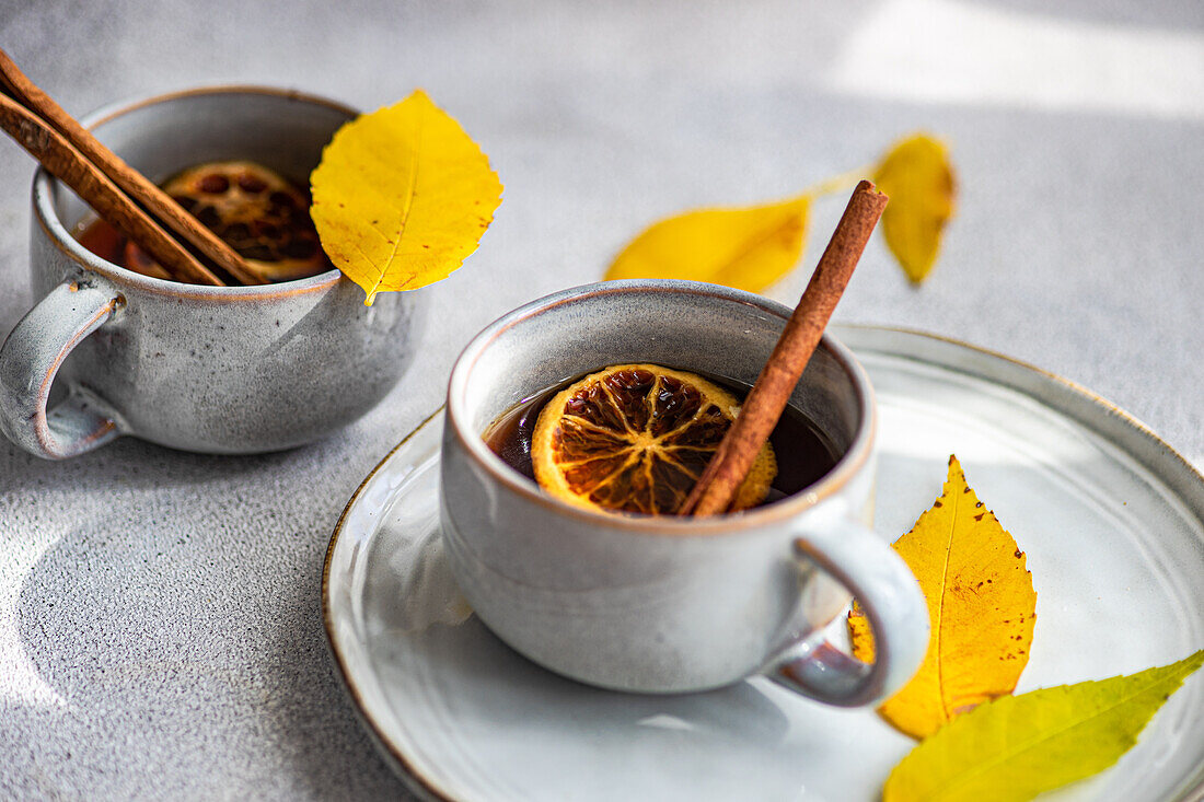 Eine warme Tasse Gewürztee, garniert mit Zimtstangen, Anis und getrockneten Orangenscheiben, ergänzt durch leuchtend gelbe, herbstliche Blätter auf einer hellgrauen Fläche