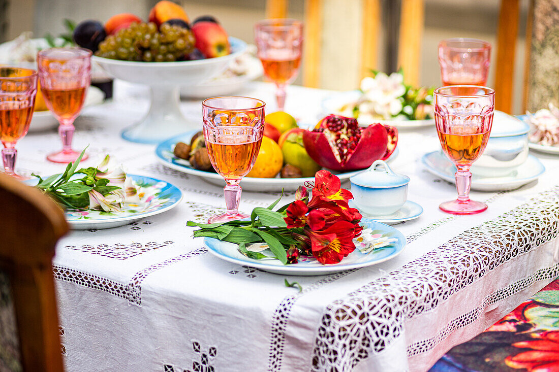 Bunt gedeckter Esstisch mit rosafarbenen Getränken in Kristallgläsern, einem Teller mit roten Blumen, verschiedenen Früchten und verziertem Porzellangeschirr, alles auf einem weißen Tischtuch mit Spitzenrand