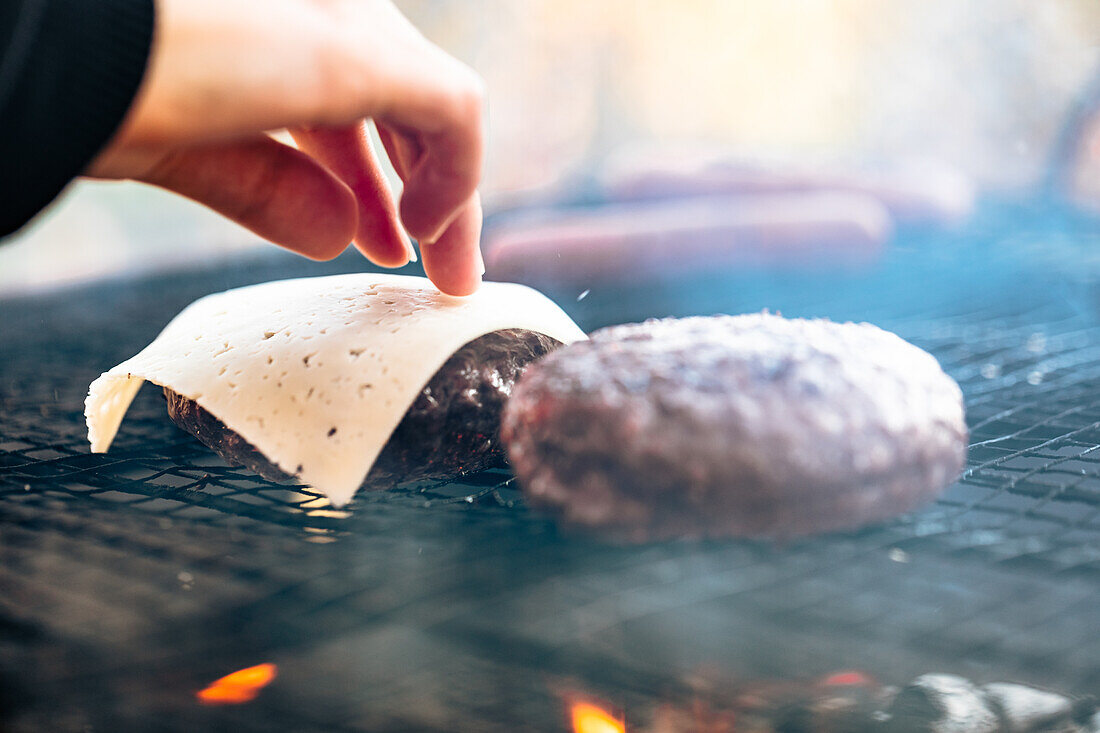 Hand placing cheese slice on burger patty over a flaming grill
