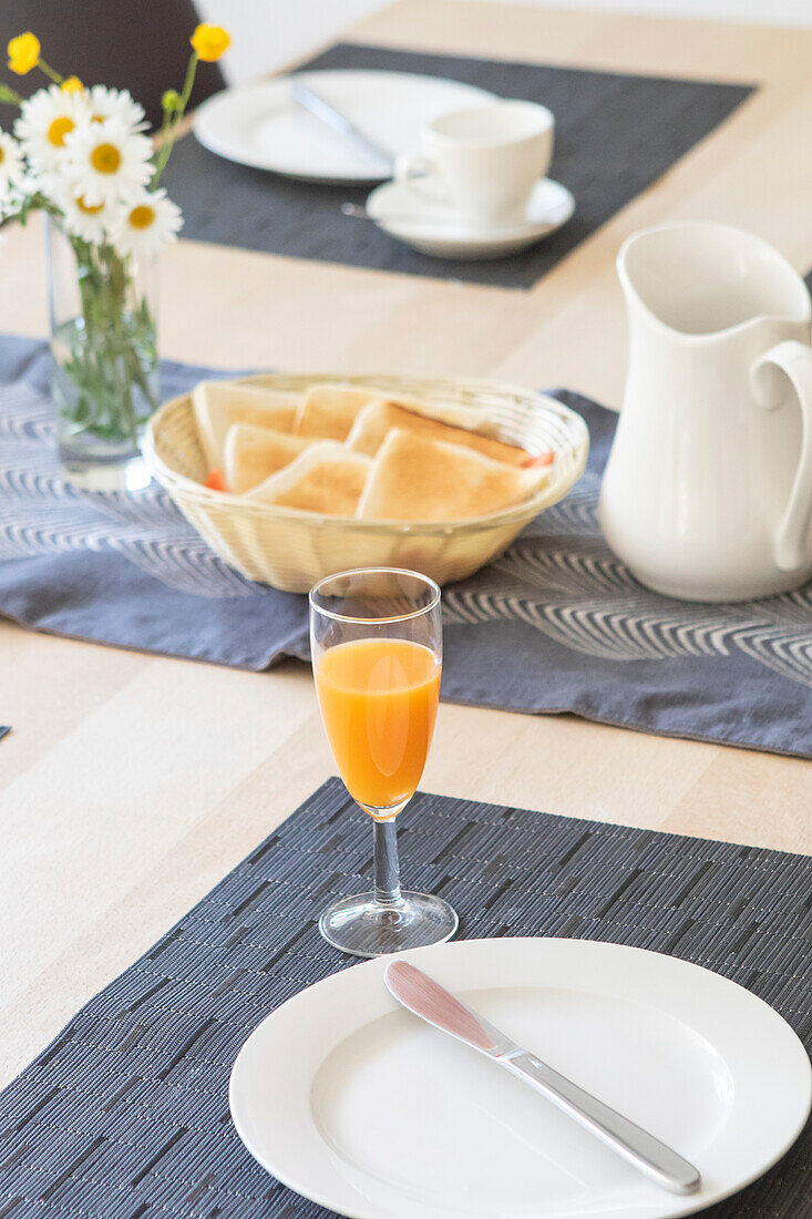Gemütliches Frühstücksambiente mit frischem Orangensaft im Glas, weißem Geschirr, einem Krug und einem geflochtenen Brotkorb, akzentuiert durch Gänseblümchen in einer Vase