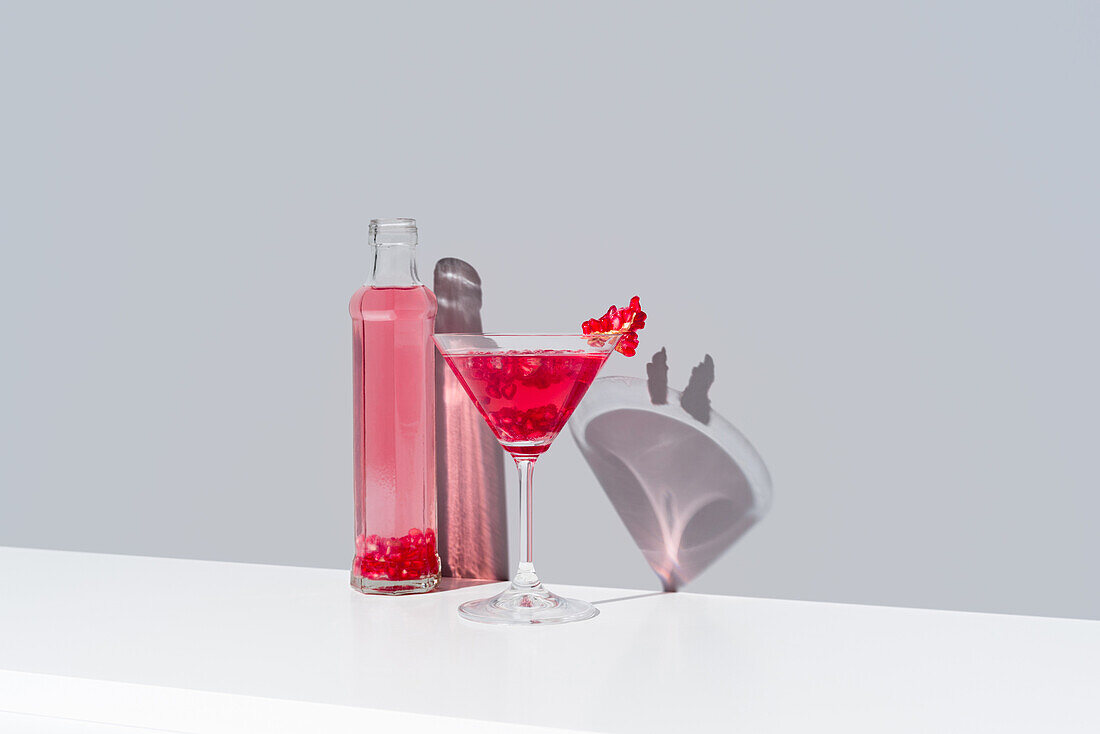 Glass filled with red pomegranate cocktail and adorned with pomegranate seeds, stands beside a transparent bottle of the same vibrant drink, both casting soft shadows on a calm gray background