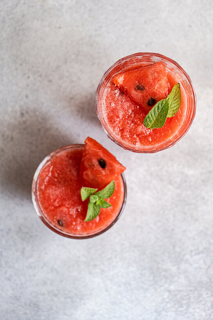 From above glasses of Margarita cocktail with watermelon smoothie garnished with leaves of mint placed on gray table