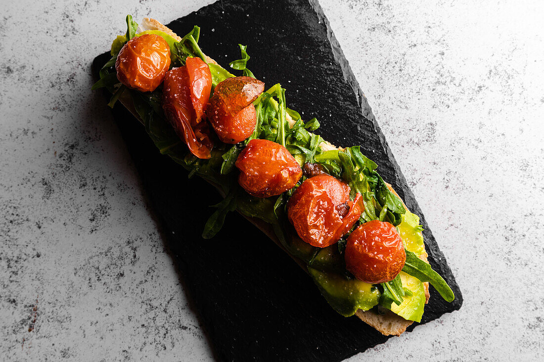 Draufsicht auf appetitliche, saftige, reife, geröstete rote Tomaten auf Avocado-Scheiben mit grünen Blättern auf schwarzer Oberfläche, serviert auf einer Marmorplatte bei Tageslicht