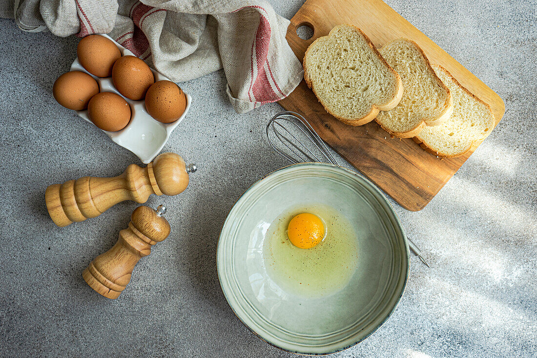Top view of raw egg in bowl near cutting board with fresh bread while cooking at concrete table with various ingredients in kitchen
