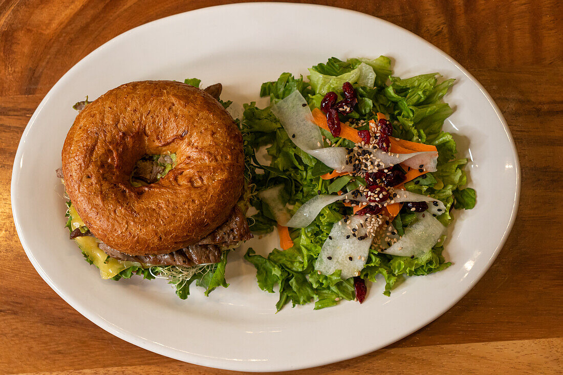 Draufsicht auf einen leckeren Burger mit Rindfleisch, Käse, frischem Salat und Karottensprossen auf einem Teller auf einer hölzernen Oberfläche im Tageslicht