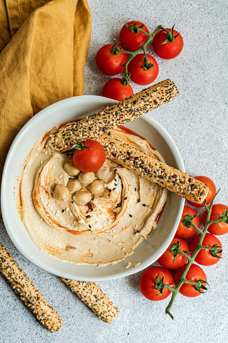 Draufsicht auf einen gesunden pflanzlichen Teller mit Hummus und Tomaten, serviert mit Brotstäbchen in einer Schüssel neben einer Serviette vor grauem Hintergrund