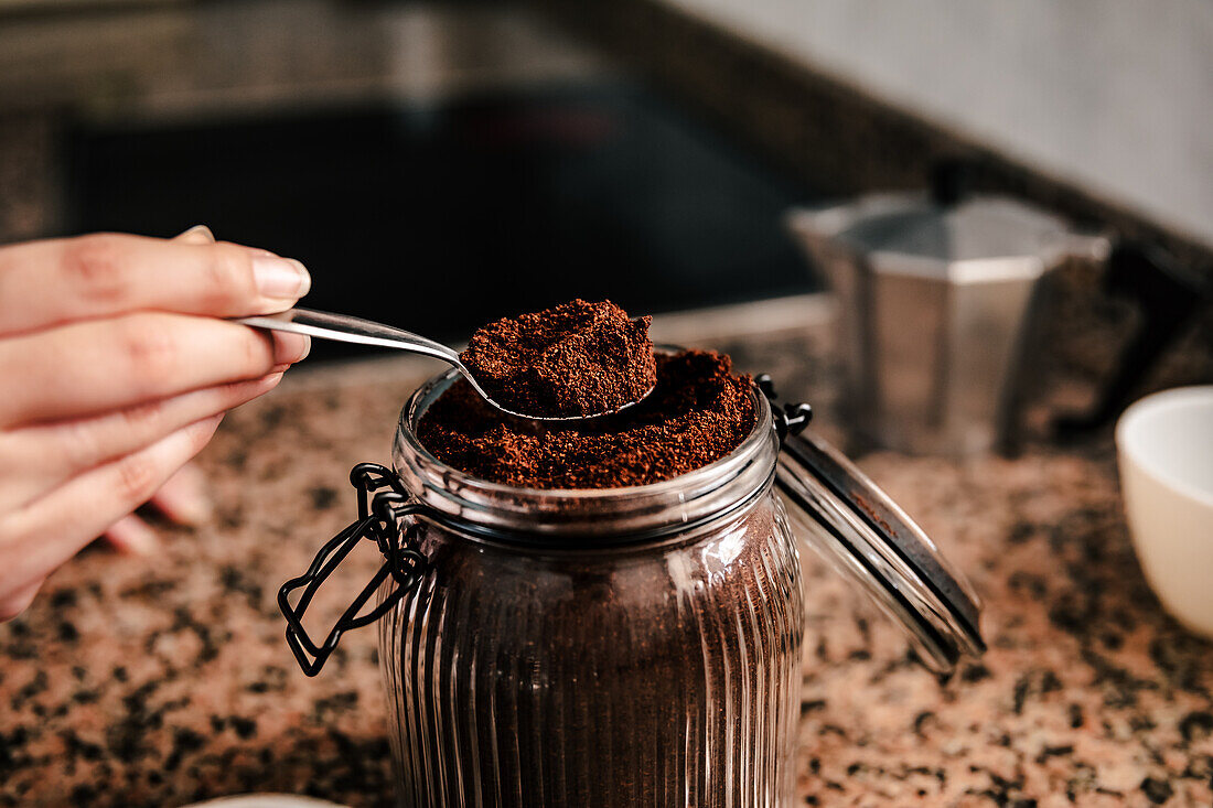 Close-up of anonymous hand scooping a spoonful of finely ground coffee from a glass jar, with a stovetop espresso maker and a cup in the background on a granite countertop
