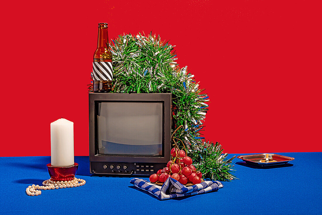 Vintage television set surrounded by an array of objects, including a bottle with a striped label, fresh grapes on a checkered cloth, a white candle, green tinsel and ashtray with cigarette butts, all set against a red backdrop on a blue table