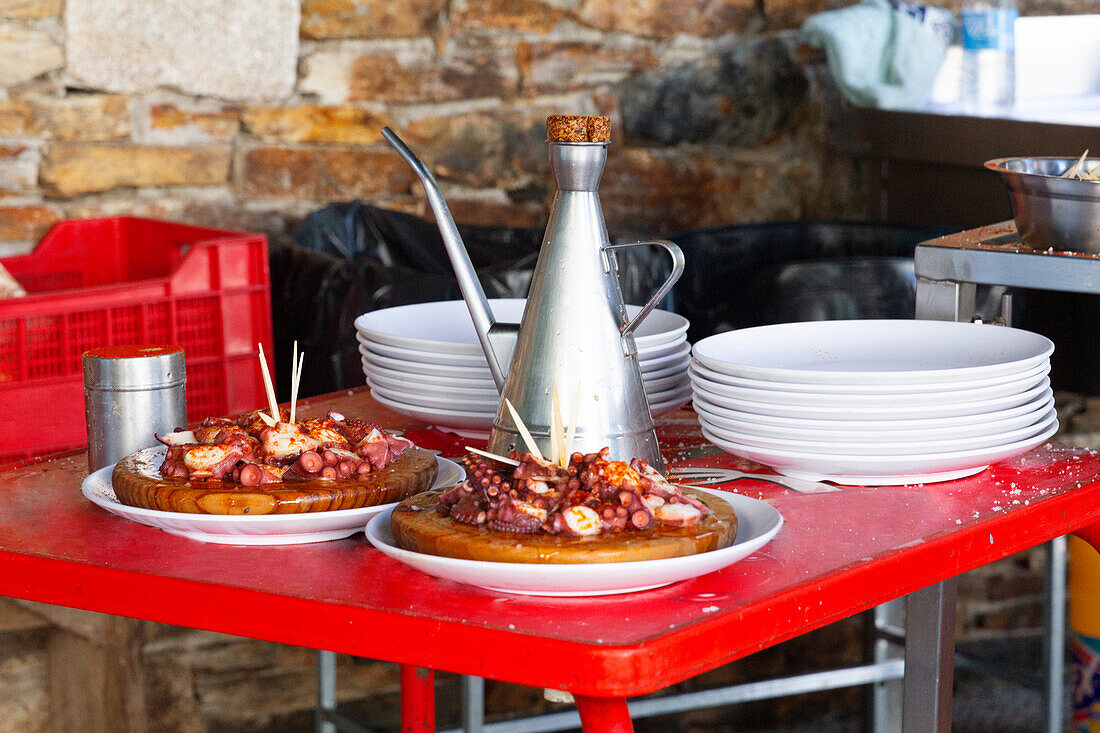 Gegrillte Oktopus-Delikatessen auf Holztellern, begleitet von einem eleganten Metallkrug, gestapelten weißen Tellern und Kochutensilien, angeordnet auf einem leuchtend roten Tisch, alles vor dem Hintergrund einer strukturierten Steinwand