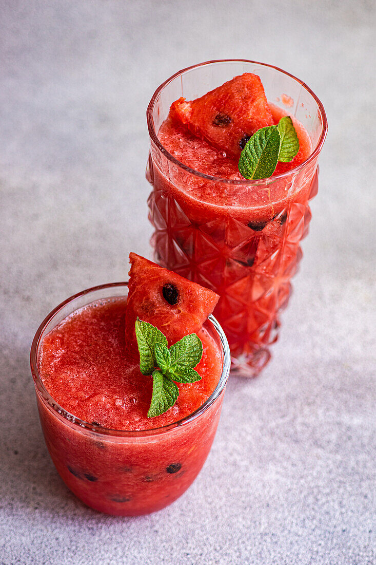 Glasses of Margarita cocktail with watermelon smoothie garnished with leaves of mint placed on gray table
