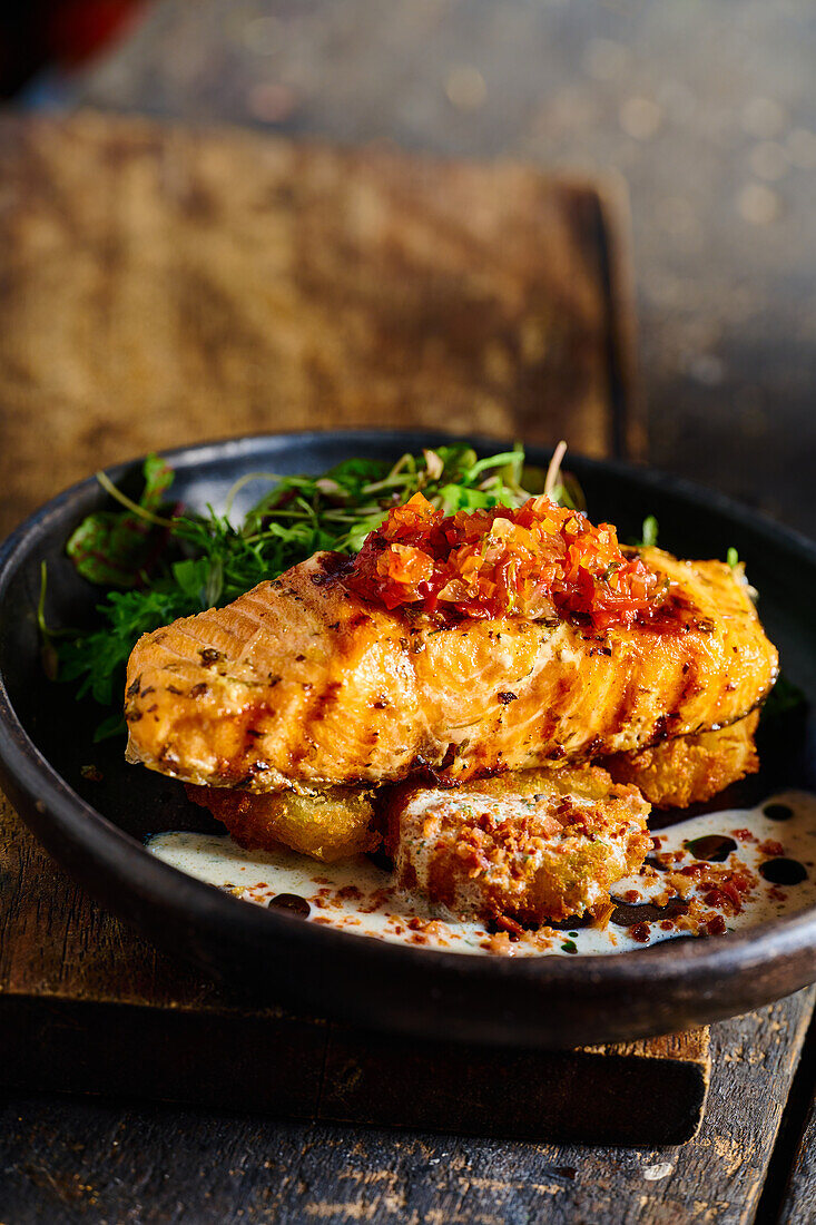 High angle of appetizing grilled salmon steak garnished with sauce and herbs served on black bowl placed on wooden cutting board