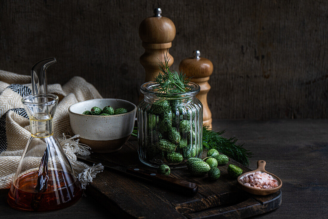 Zutaten für die Zubereitung von fermentierten Gurken in einem Glas auf einem Holztablett neben einer Serviette und Salz- und Pfefferstreuern vor dunklem Hintergrund