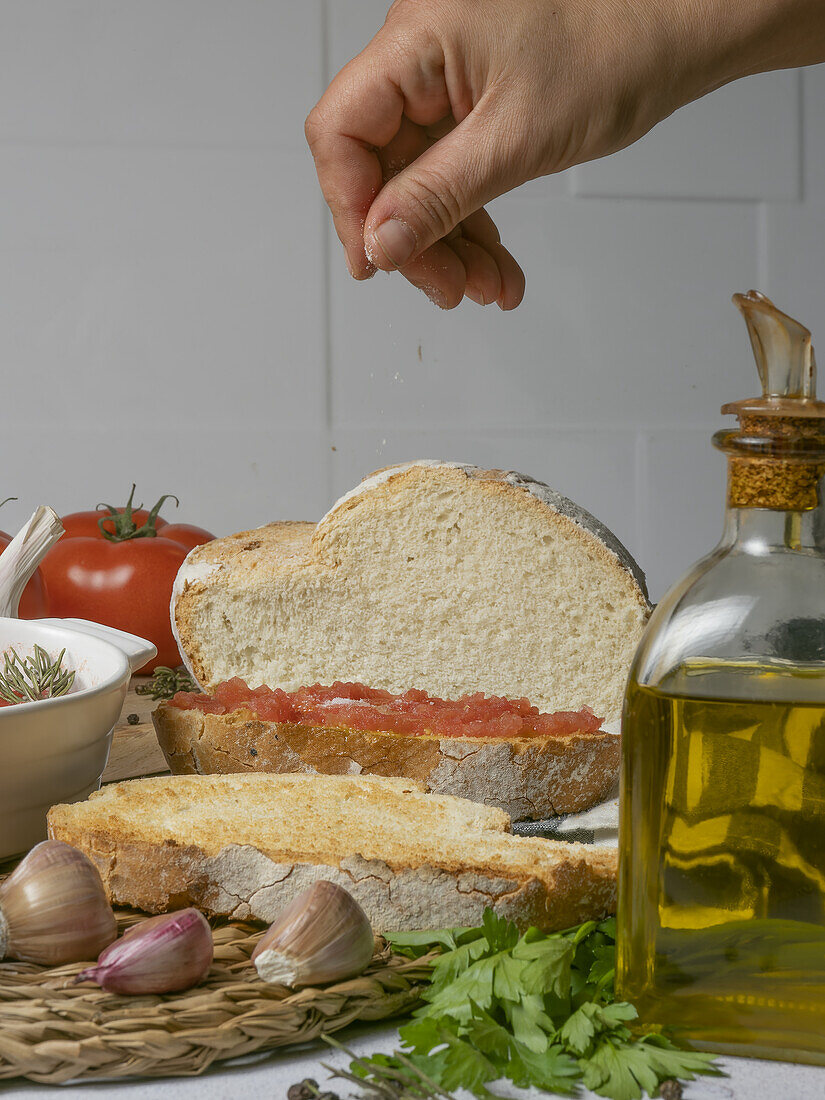 Crop hand of anonymous cook sprinkling salt on bread with tomato spread placed on table against bottle of olive oil and unpeeled garlic with greens