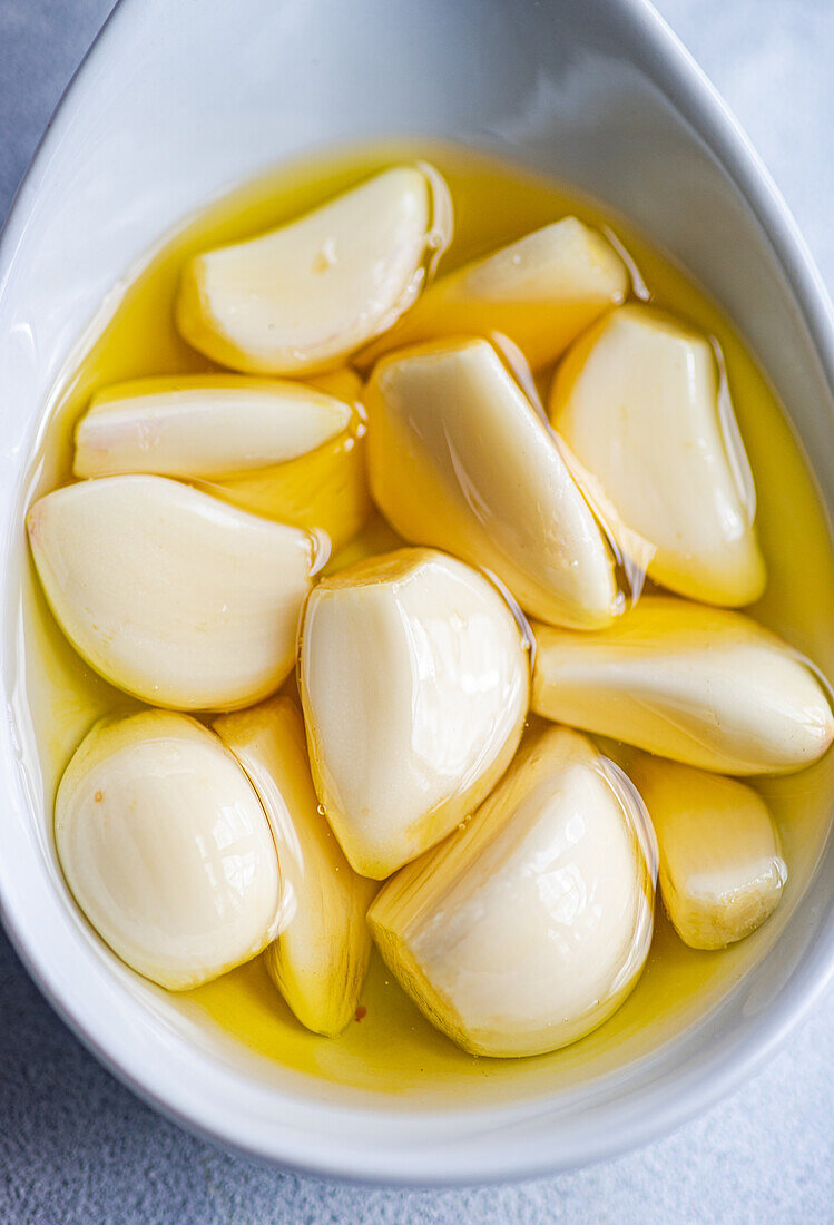 Top view of closeup peeled and baked garlic cloves submerged in oil, showcased in a ceramic spoon against a light gray background