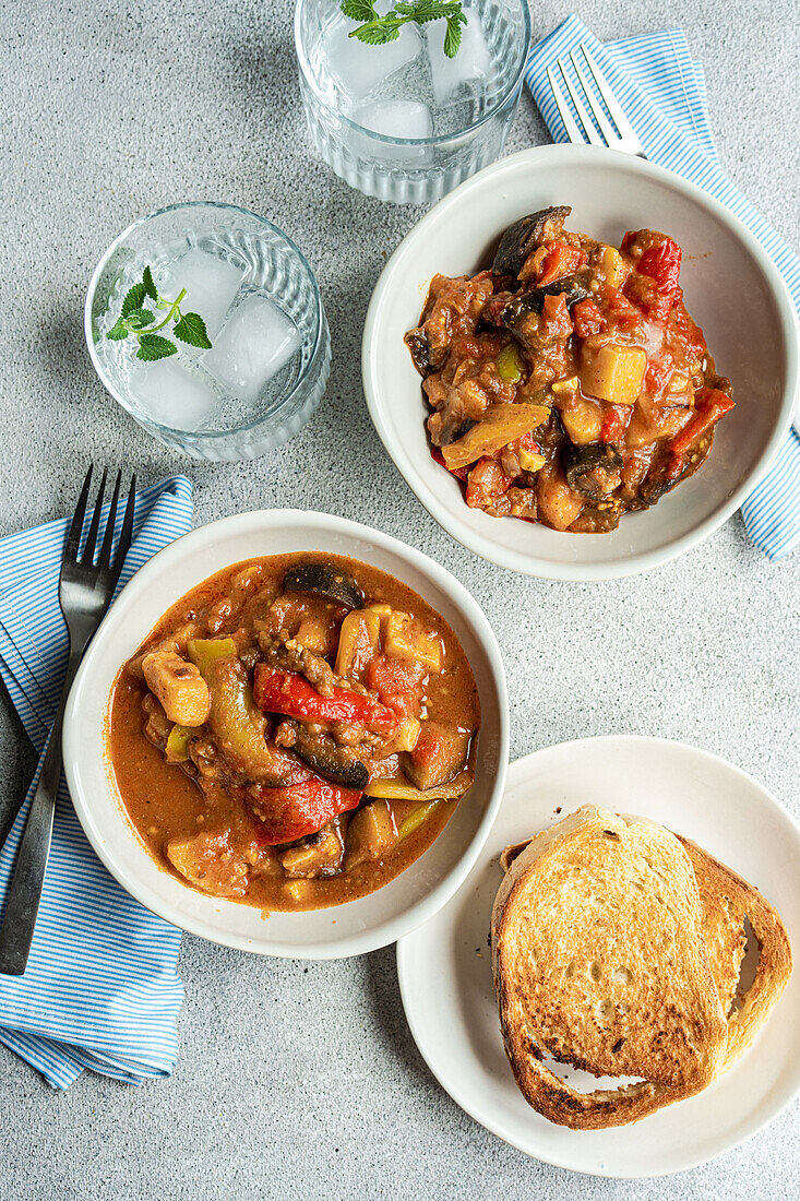 Top view of traditional seasonal Georgian dish - Adjapsandali with stewed vegetables like potato, bell peppers, tomato, egg plant, coriander and Georgian spices placed on gray table near napkin and glass with water