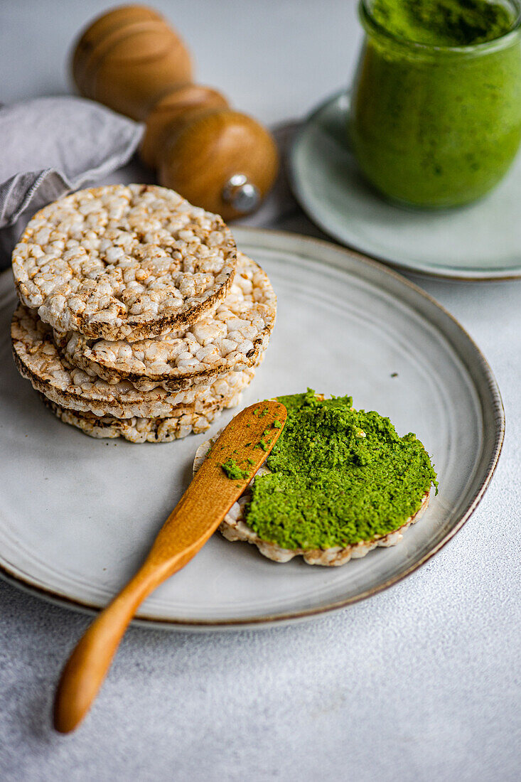 High angle of rice bread on a plate accompanied by a vibrant green spinach pesto pasta-sauce in a glass jar, set against a blurred backdrop near napkin and salt shaker