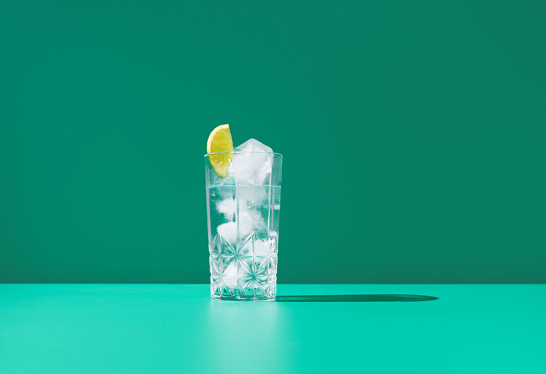 A clear glass full of gin tonic and topped with a lime slice sits against a vibrant green backdrop, creating a fresh and cool visual.