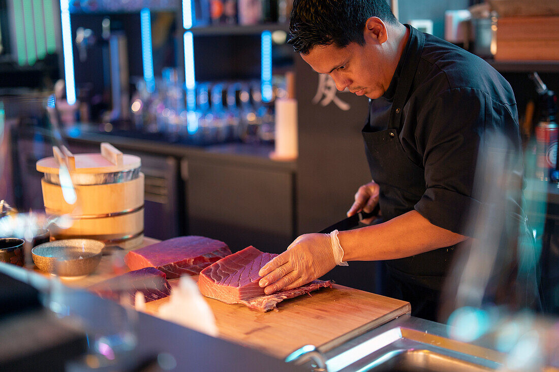 A sushi chef attentively slices fresh fish with a sharp knife at a wooden counter in the ambient lighting of a modern sushi restaurant.