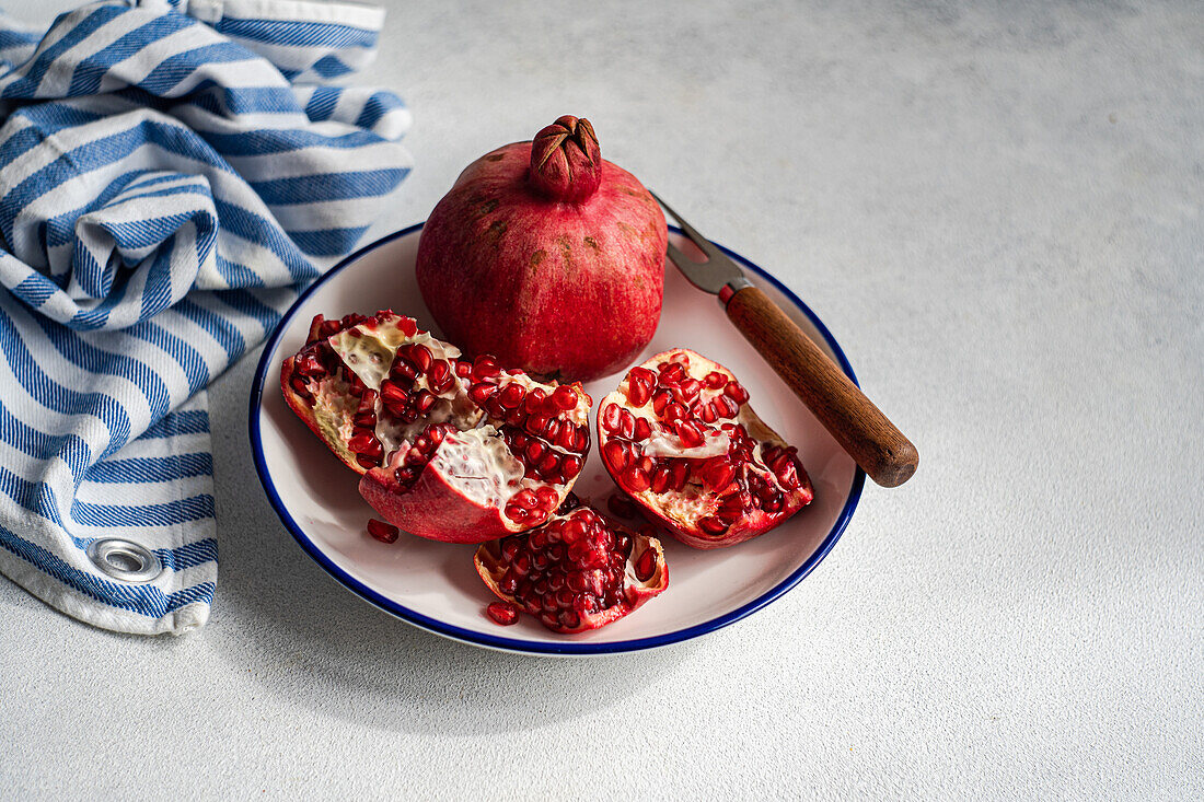 A fresh ripe pomegranate sits on a white and blue plate, surrounded by its red seeds and a rustic knife. A striped napkin blue and white against gray background.