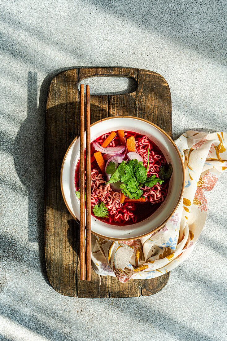 Schale mit Rote-Bete-Suppe mit Zwiebeln, Koriander und Nudeln im asiatischen Stil, serviert auf einer Schale und Stäbchen auf einer Serviette vor einer grauen Fläche im Tageslicht (Draufsicht)