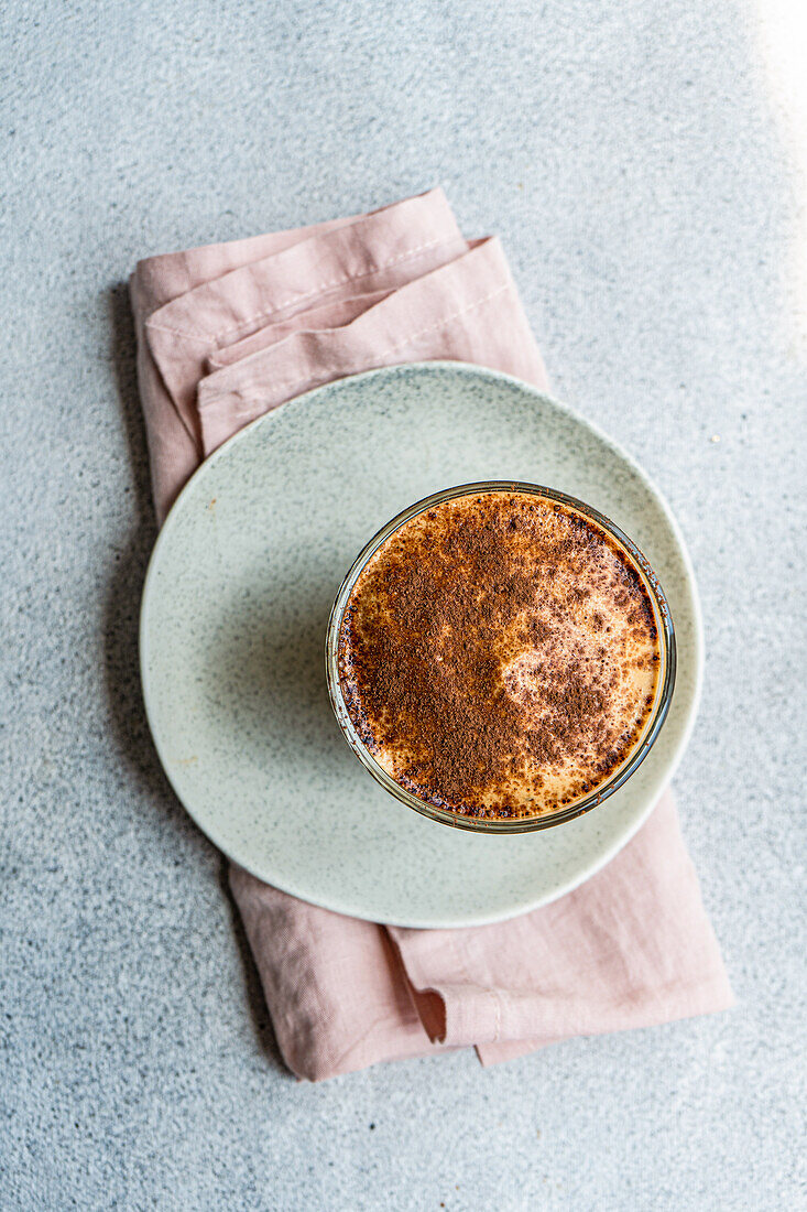 Draufsicht auf eine Glastasse mit Kaffeesahne-Dessert mit Schokoladenpulver und Eis, serviert auf einem Teller mit Serviette für eine erfrischende Sommermahlzeit vor verschwommenem Hintergrund