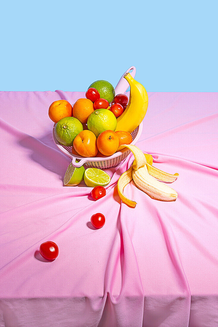 Plastic bowl with fresh assorted fruits placed on at table with a pink tablecloth table in daylight on blue background