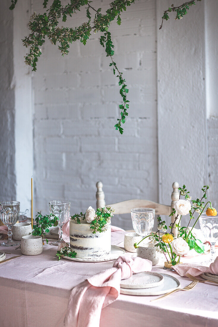Eleganter Tisch mit Tellern und Blumen in der Nähe eines leckeren Kuchens auf einem rosa Tischtuch vor einer Backsteinwand
