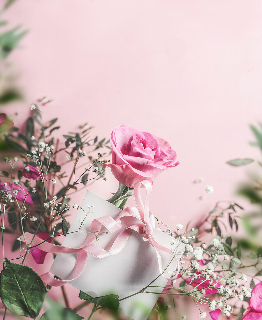 White greeting card with ribbon and rose flowers at pink background with green leaves frame. Top view with copy space.