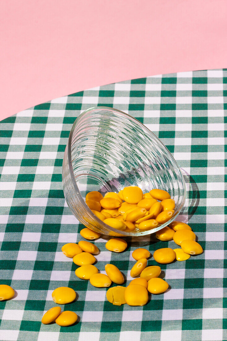 Transparente Glasschale mit gelben Lupinenbohnen auf einem karierten Tisch vor rosa Hintergrund