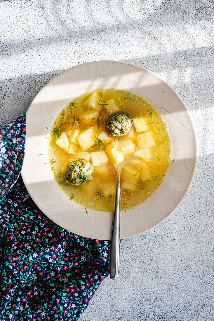 From above sunlit bowl of homemade meatball soup with diced potatoes, on a textile surface.
