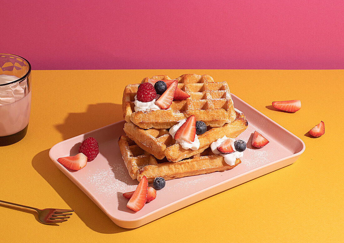 From above tasty sweet waffles topped with berries fruits sauce and cream served on pink tray on colorful table background near glass with hot milk beverage in light kitchen