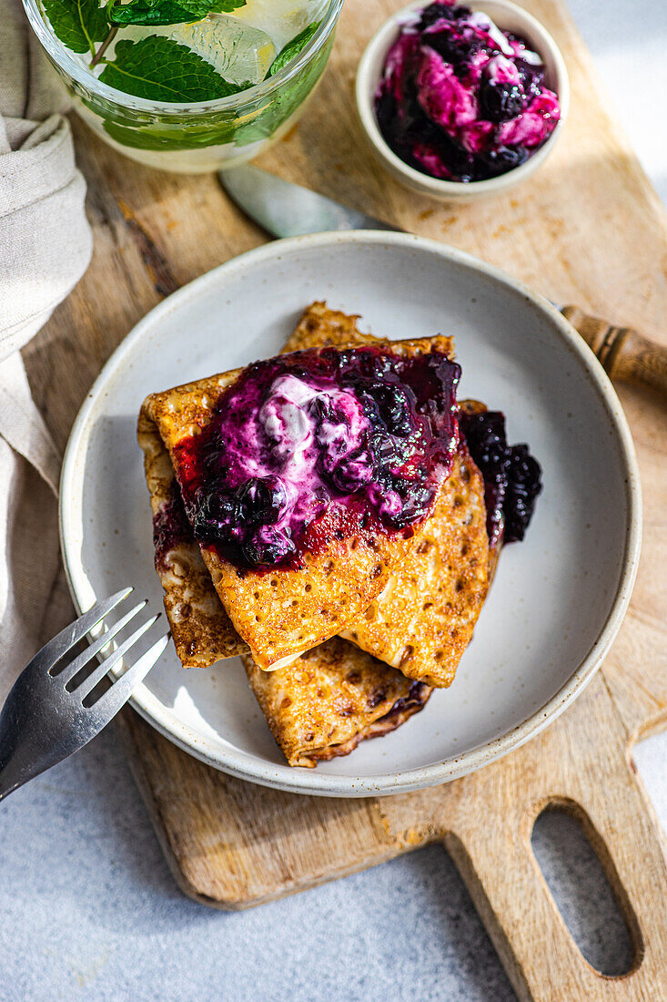 Golden crepes topped with blueberry jam and a dollop of sour cream, served on a rustic wooden board.