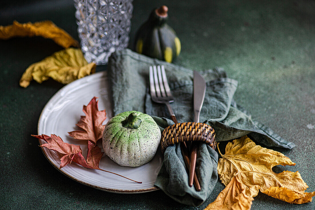 High angle of autumnal table setting with napkin, knife and fork place on plate near colorful leaves, smalls pumpkins and empty glass against dark surface