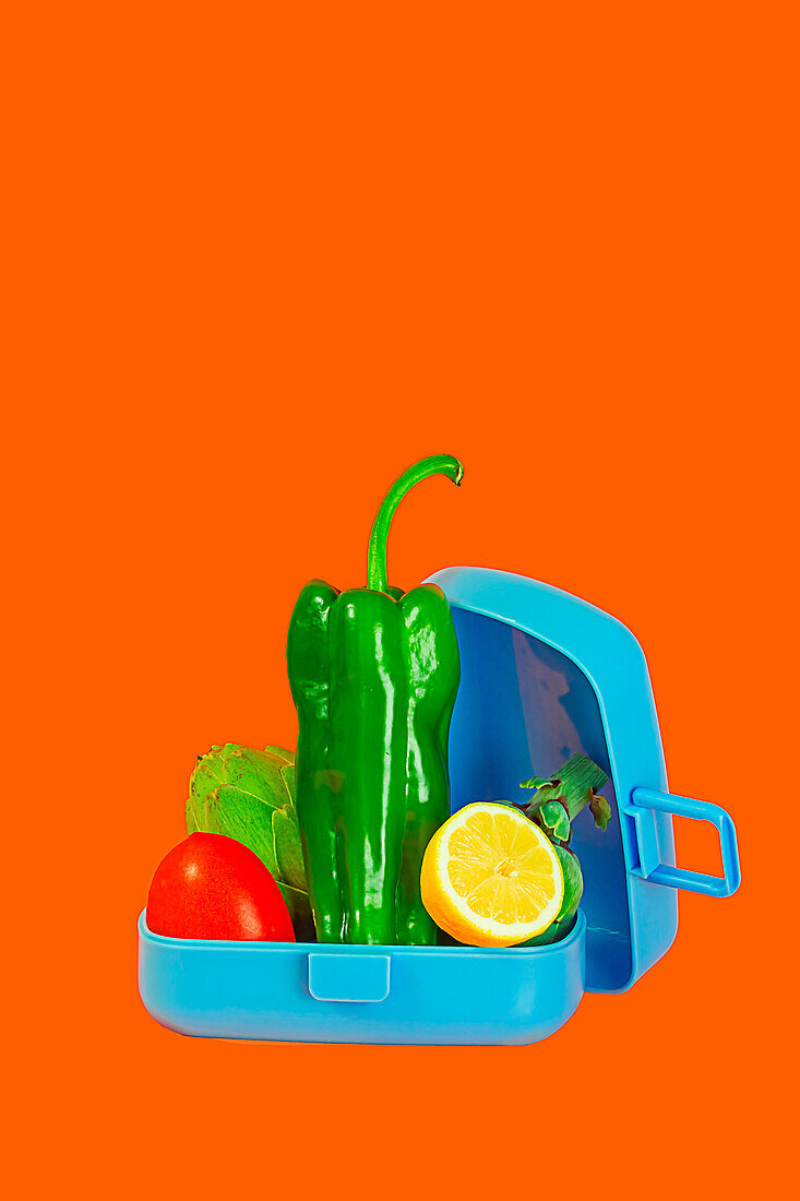 Eine offene Lunchbox, gefüllt mit einer Vielzahl von frischem Gemüse, darunter Paprika und Zitrone, vor einem leuchtend orangefarbenen Hintergrund
