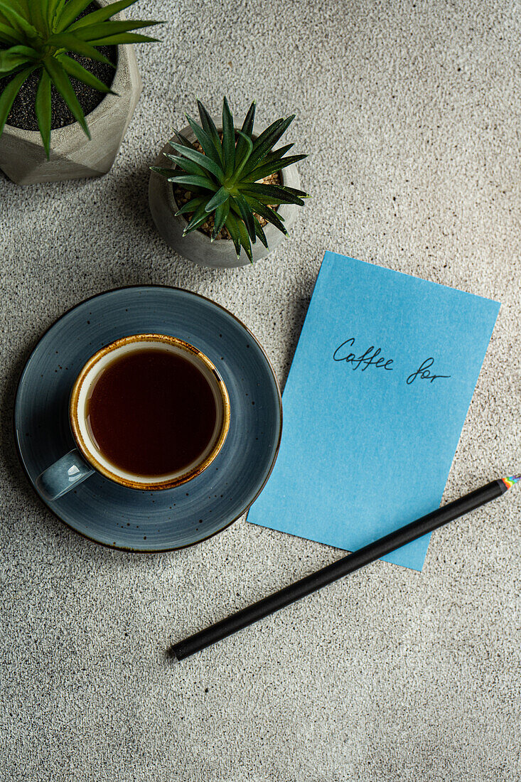 Draufsicht auf eine Tasse Kaffee neben Topfpflanzen und einer Notiz mit Bleistift auf einem grauen Tisch