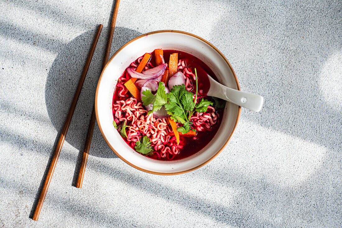 Top view of bowl with beetroot soup with onion, coriander and noodles in Asian style served on bowl with spoon and chopsticks against gray surface in daylight