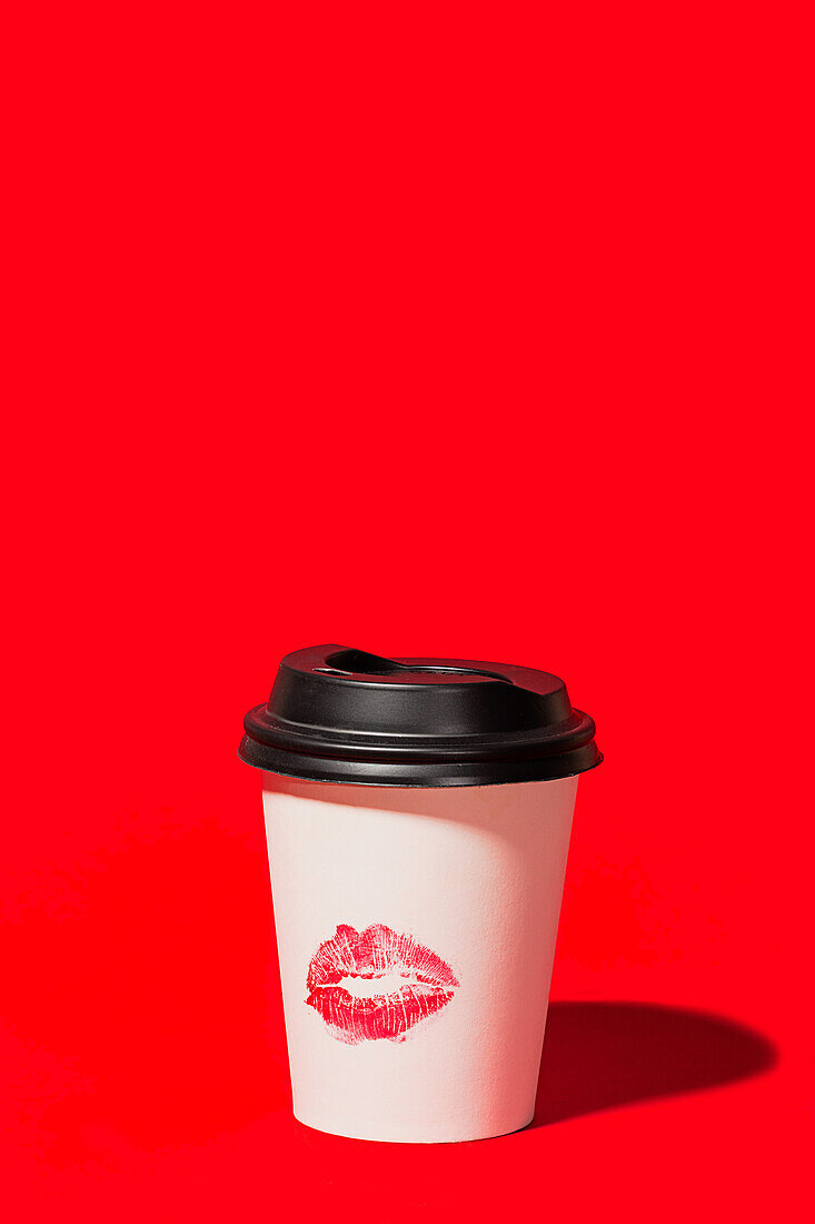 Ein einzelner weißer Einweg-Kaffeebecher mit schwarzem Deckel und einem roten Lippenstift-Kuss-Zeichen steht auf einem lebhaften roten Hintergrund, der eine kühne und romantische Aussage macht