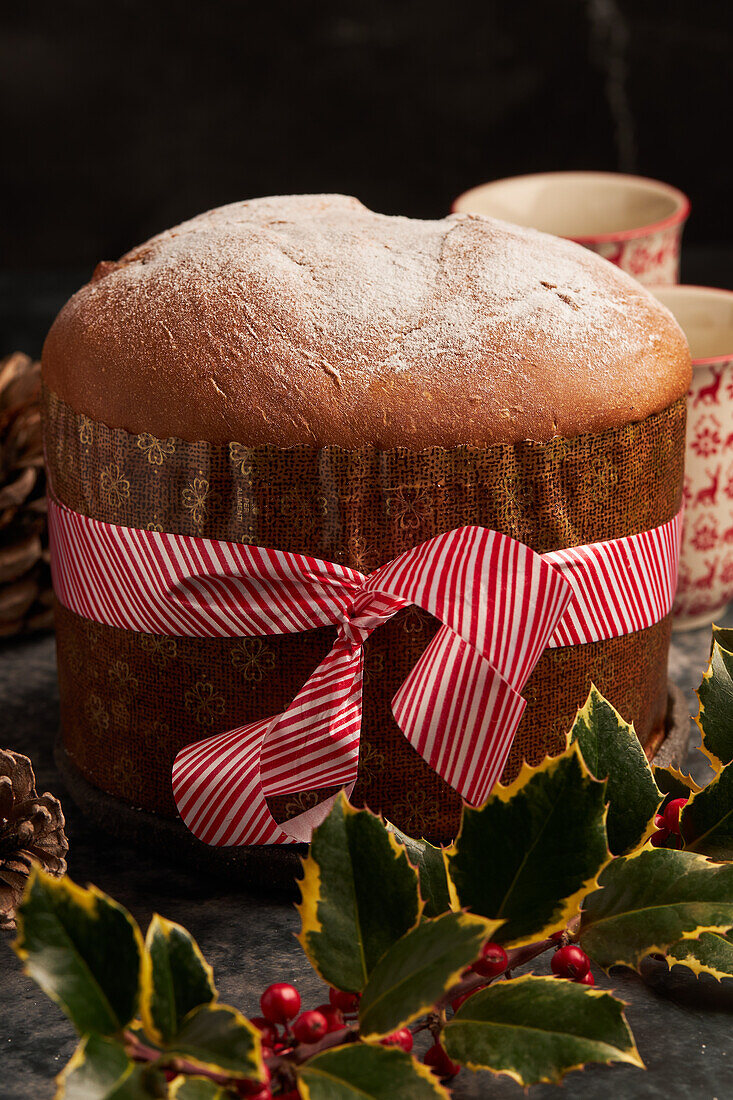 A traditional panettone dusted with icing sugar, adorned with a decorative ribbon, surrounded by holly leaves and berries, evoking the warmth of the holiday season