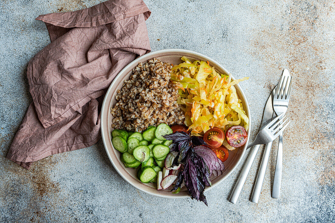 Top view of bowl with delicious buckwheat containing sliced fresh basil plant with cabbage and cucumber with cutlery placed on gray table with napkin